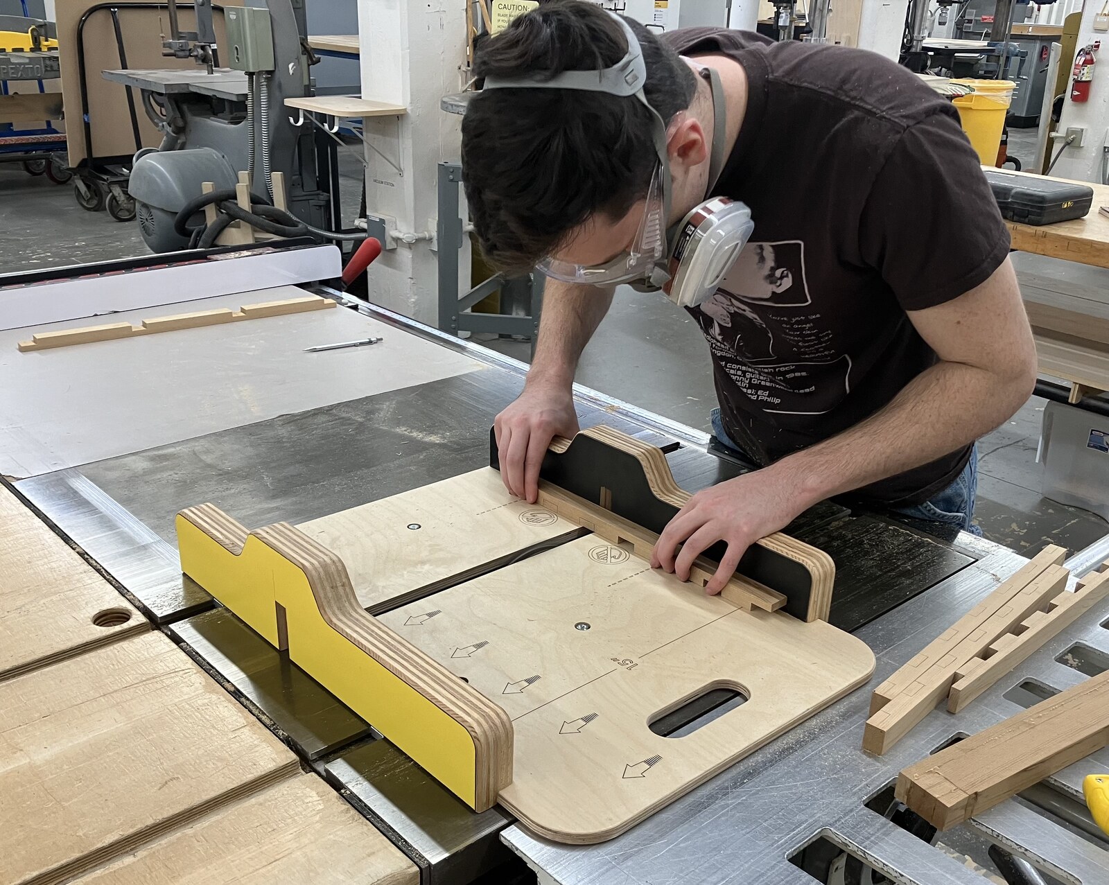 George at a table saw in the wood shop, guiding a workpiece through a shop-built jig.